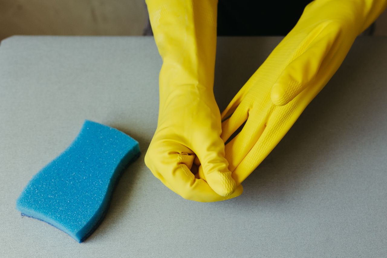 services-01 Close-up of hands wearing yellow gloves next to blue sponge on gray surface.